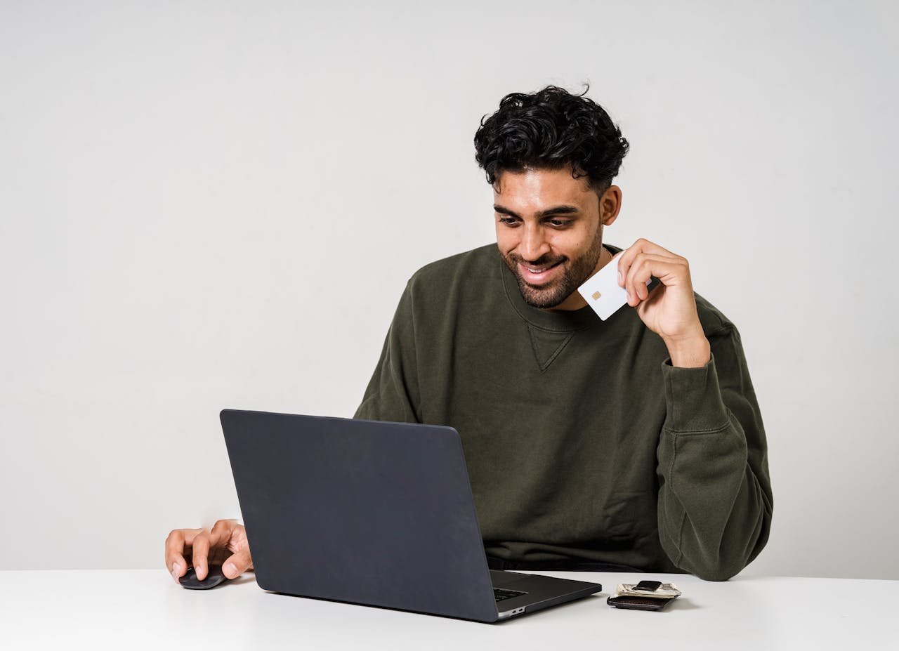 A man smiling while using a laptop and holding a credit card for online shopping.