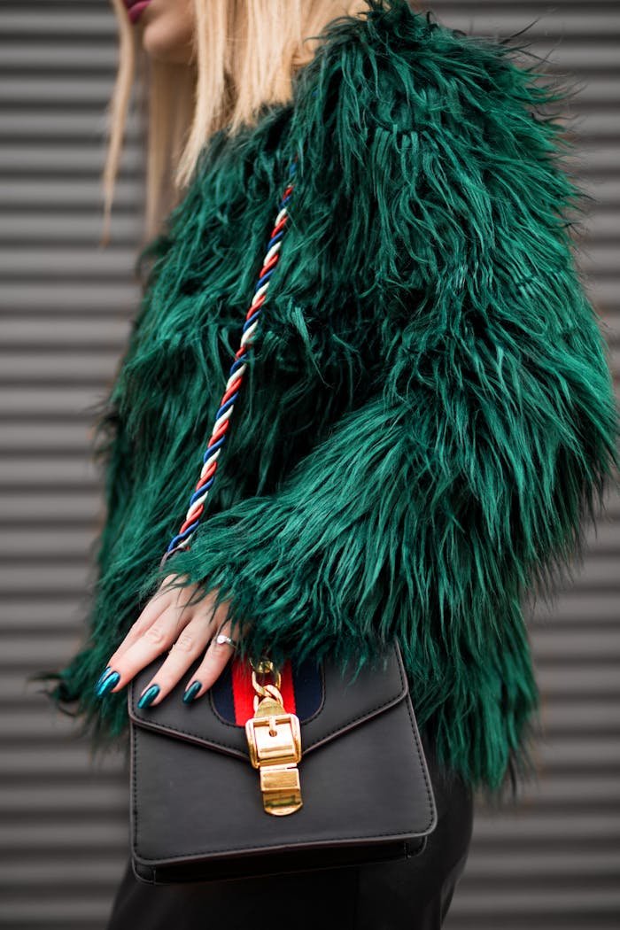 brand-03 Stylish woman in a green faux fur jacket holding a fashionable handbag outdoors.