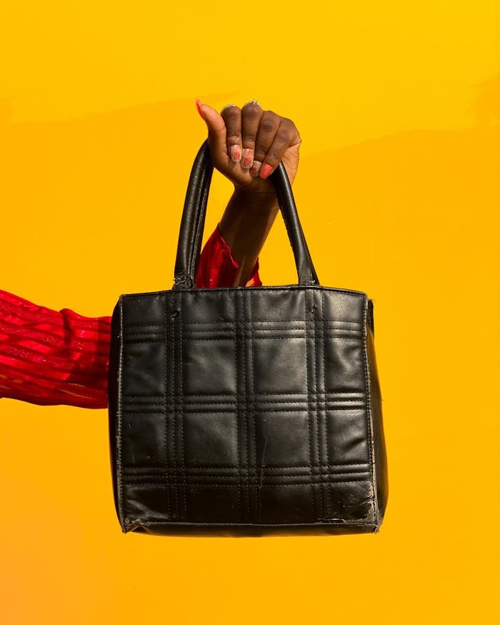 A fashionable black handbag is held by a hand with a vibrant yellow background, showcasing modern style.