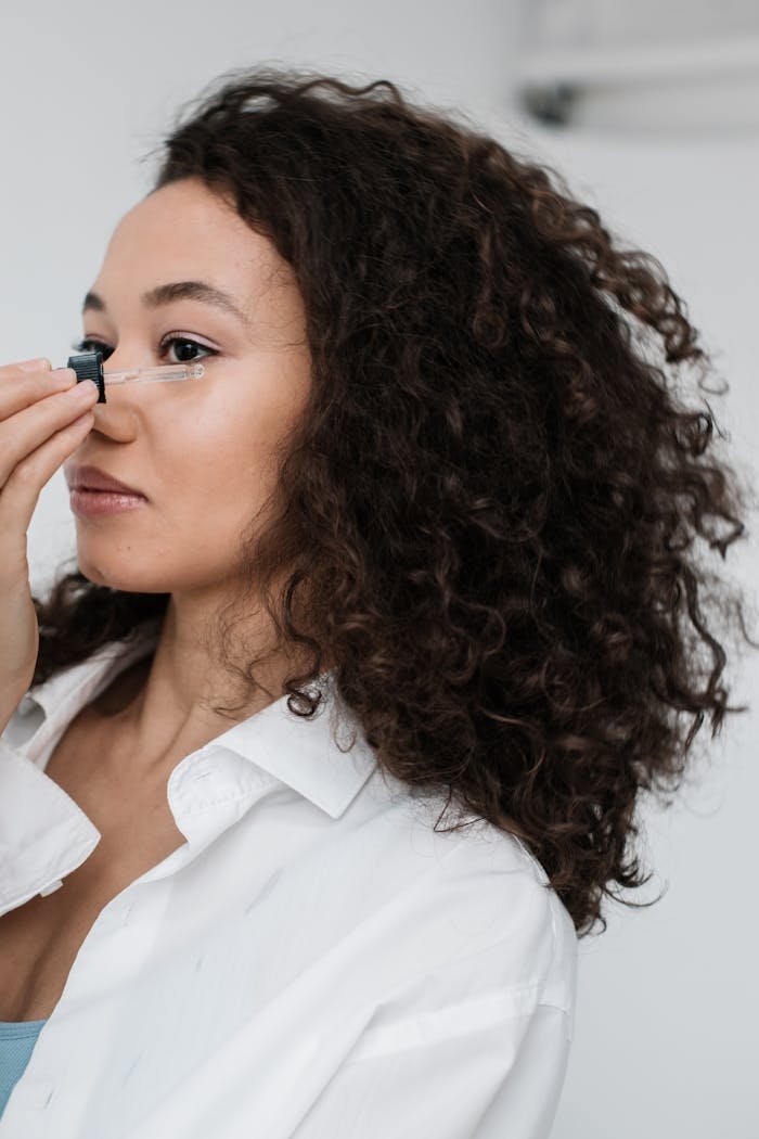 mobile-02 Woman with curly hair applying skincare oil to her face, focusing on self-care and beauty routine.