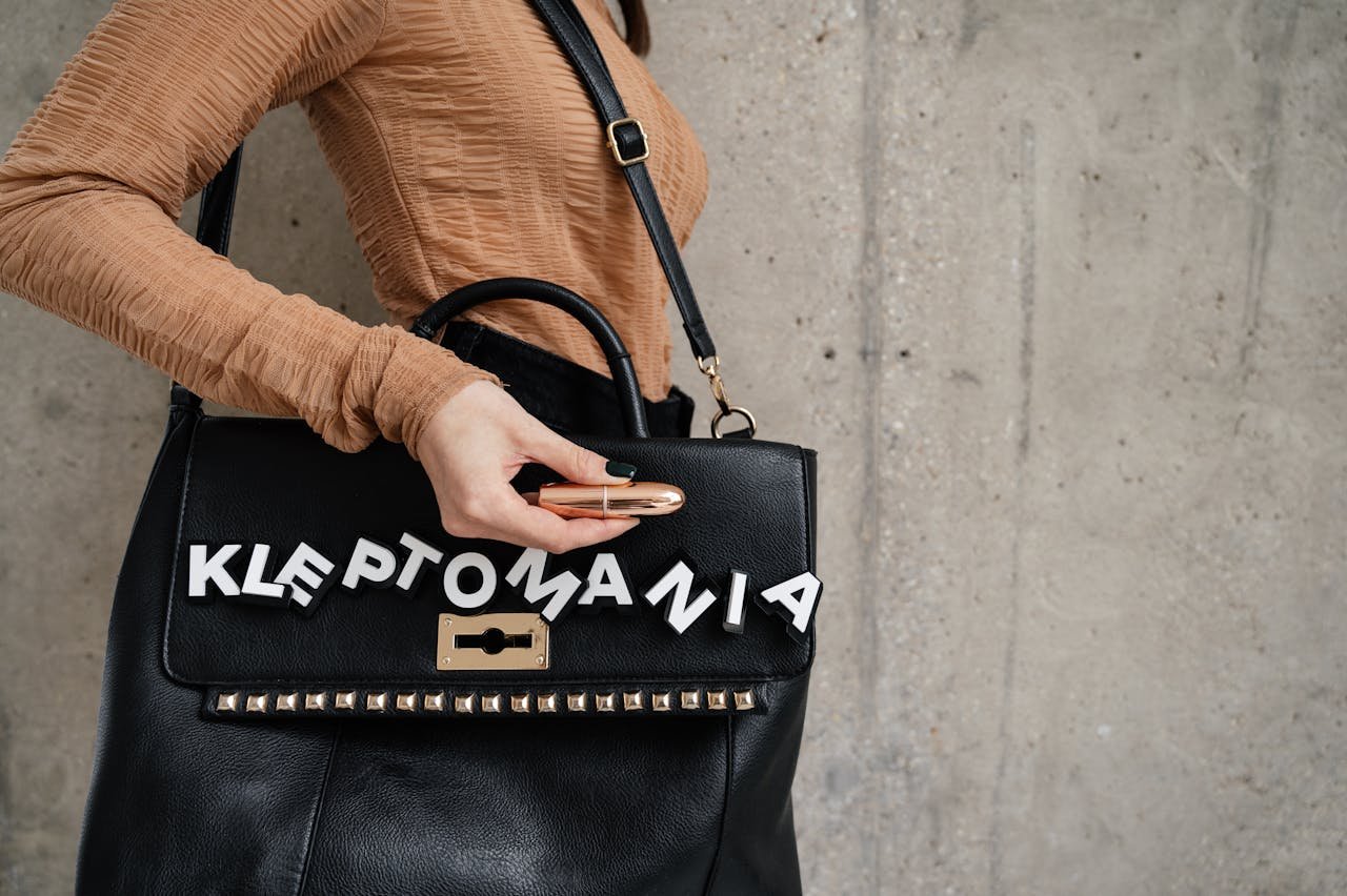 our-story Close-up of a black handbag with 'Kleptomania' letters and a lipstick in hand.