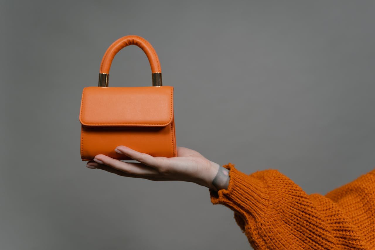 brand-02 Stylish orange handbag held by a hand against a gray background. Perfect for modern fashion.